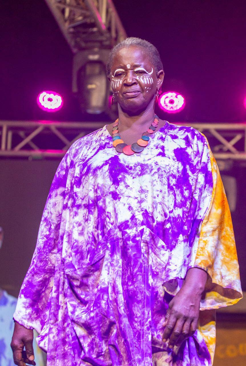 Une mannequin défile dans une robe violette sous les lumières de la scène.